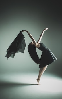 A dancer gracefully arches backward, balancing on one foot with long hair cascading down. She holds a flowing black fabric in one hand, creating a sense of motion. The background is softly lit, with subtle shadows accentuating her form.