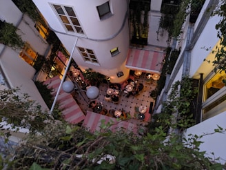 Aerial view of an outdoor restaurant courtyard surrounded by tall buildings with ivy-covered walls. The courtyard features checkered flooring and multiple round tables with people dining. The area is illuminated by warm, glowing lights enhancing the cozy atmosphere.