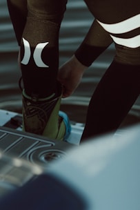 A person is putting on or adjusting water sports gear, standing on a platform above a body of water. The lower part of their body is visible, dressed in a black wetsuit with prominent white logos. Their footwear appears specialized, suggesting activities like wakeboarding or water skiing.