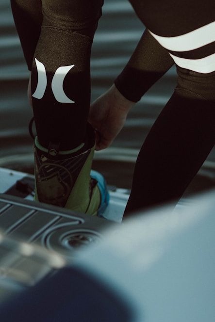 A person is putting on or adjusting water sports gear, standing on a platform above a body of water. The lower part of their body is visible, dressed in a black wetsuit with prominent white logos. Their footwear appears specialized, suggesting activities like wakeboarding or water skiing.