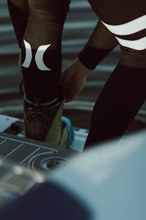 A person is putting on or adjusting water sports gear, standing on a platform above a body of water. The lower part of their body is visible, dressed in a black wetsuit with prominent white logos. Their footwear appears specialized, suggesting activities like wakeboarding or water skiing.
