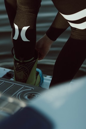 A person is putting on or adjusting water sports gear, standing on a platform above a body of water. The lower part of their body is visible, dressed in a black wetsuit with prominent white logos. Their footwear appears specialized, suggesting activities like wakeboarding or water skiing.