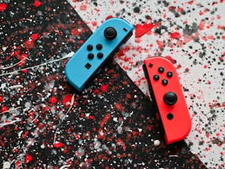 Two video game controllers, one blue and one red, are placed on a splattered paint background with black, red, and white colors.
