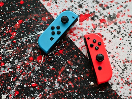 Two video game controllers, one blue and one red, are placed on a splattered paint background with black, red, and white colors.