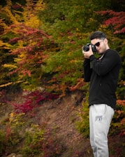 A person is taking a photograph with a digital camera, standing amidst vibrant autumn foliage. They are dressed in casual attire, including a black jacket and gray sweatpants.