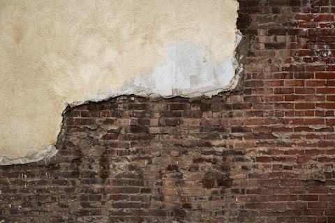 A weathered brick wall with patches of peeling plaster and exposed masonry, showcasing a blend of rough and smooth textures. The plaster has worn away in several areas, revealing the uneven and aged bricks beneath.