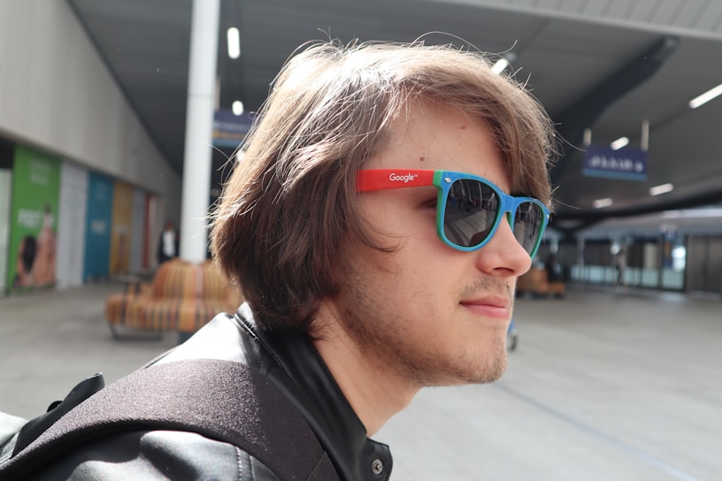 A person with shoulder-length hair wearing vibrant sunglasses and a black backpack, standing in a spacious, modern indoor setting. The sunglasses have a red and blue frame with a logo on the side. The background features a high ceiling, benches, and a few colorful posters on the walls.
