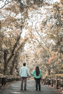 A couple holding hands walks along a tree-lined path in a serene forest setting. The pathway is flanked by wooden railings, and the tall trees overhead create a canopy of leaves, filtering the light. The overall scene is tranquil and lush, with a mix of green and brown tones.