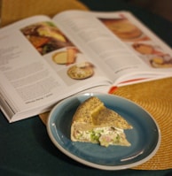 A slice of pie with a cream filling, flecked with herbs and pieces of ham, is on a blue plate. Behind it, an open cookbook displays various recipes and images on a woven mat.