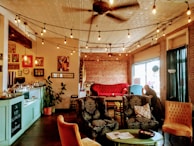 A cozy cafe interior featuring a variety of vintage and upholstered furniture, including a red velvet sofa and ornamental chairs. The walls are adorned with exposed brick and eclectic artwork. Soft string lights hang from a ceiling fan, creating a warm and inviting atmosphere. A man sits by the window, adding to the relaxing ambiance of the space.