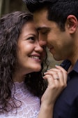 A couple is captured in a tender moment, with their faces close and smiling affectionately. The woman has curly hair and is wearing a white lace top, and a ring is visible on her left hand. The man has short dark hair and is wearing a dark shirt. Their expressions convey warmth and closeness.