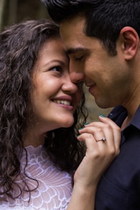 A couple is captured in a tender moment, with their faces close and smiling affectionately. The woman has curly hair and is wearing a white lace top, and a ring is visible on her left hand. The man has short dark hair and is wearing a dark shirt. Their expressions convey warmth and closeness.