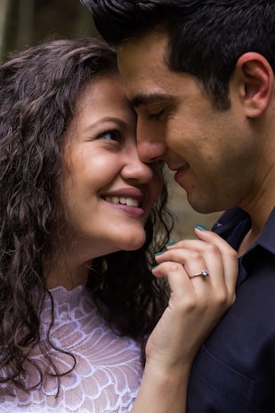 A couple is captured in a tender moment, with their faces close and smiling affectionately. The woman has curly hair and is wearing a white lace top, and a ring is visible on her left hand. The man has short dark hair and is wearing a dark shirt. Their expressions convey warmth and closeness.