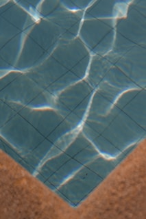 A section of a swimming pool with clear blue water, showing the grid pattern of the pool tiles. Sunlight casts bright, wavy reflections across the water's surface. The image features the pool’s corner with a contrasting tan edge.