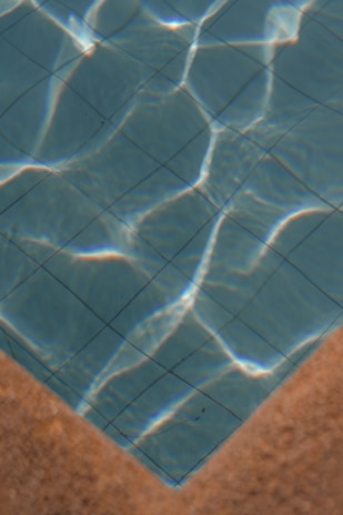 A section of a swimming pool with clear blue water, showing the grid pattern of the pool tiles. Sunlight casts bright, wavy reflections across the water's surface. The image features the pool’s corner with a contrasting tan edge.