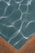 A section of a swimming pool with clear blue water, showing the grid pattern of the pool tiles. Sunlight casts bright, wavy reflections across the water's surface. The image features the pool’s corner with a contrasting tan edge.