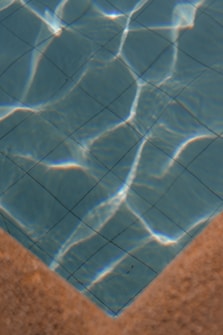 A section of a swimming pool with clear blue water, showing the grid pattern of the pool tiles. Sunlight casts bright, wavy reflections across the water's surface. The image features the pool&rsquo;s corner with a contrasting tan edge.