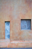 A minimalistic exterior wall features a small, rectangular window and a narrow vertical door. The wall has a textured surface, predominantly in earthy tones. The window shows signs of wear with dust or residue on the glass, while the door has circular patterns on a metallic surface.