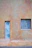 A minimalistic exterior wall features a small, rectangular window and a narrow vertical door. The wall has a textured surface, predominantly in earthy tones. The window shows signs of wear with dust or residue on the glass, while the door has circular patterns on a metallic surface.