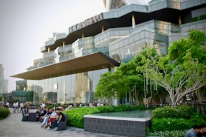 A modern retail building with a distinctive architectural design, featuring large glass panels and a prominently displayed logo of a well-known technology company. The surrounding area includes well-maintained greenery, trees, and several people sitting and walking nearby, suggesting a lively yet relaxed atmosphere.