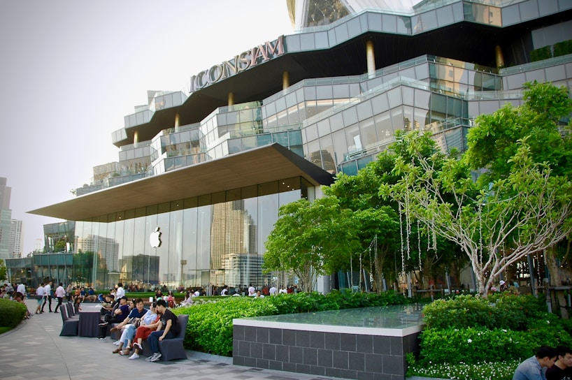 A modern retail building with a distinctive architectural design, featuring large glass panels and a prominently displayed logo of a well-known technology company. The surrounding area includes well-maintained greenery, trees, and several people sitting and walking nearby, suggesting a lively yet relaxed atmosphere.