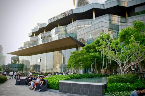 A modern retail building with a distinctive architectural design, featuring large glass panels and a prominently displayed logo of a well-known technology company. The surrounding area includes well-maintained greenery, trees, and several people sitting and walking nearby, suggesting a lively yet relaxed atmosphere.