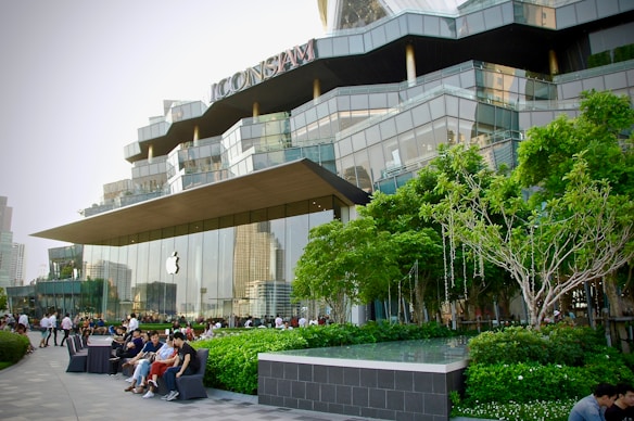 A modern retail building with a distinctive architectural design, featuring large glass panels and a prominently displayed logo of a well-known technology company. The surrounding area includes well-maintained greenery, trees, and several people sitting and walking nearby, suggesting a lively yet relaxed atmosphere.