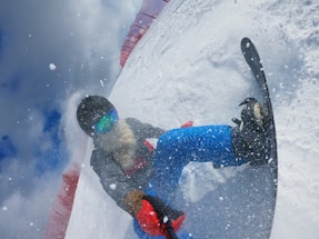 A snowboarder is captured during an intense action moment on a snowy slope. The person is wearing a helmet, colorful reflective goggles, and winter attire including a grey jacket and blue pants. Snow is flying around, indicating speed and movement, and a red snow barrier is visible along the slope.