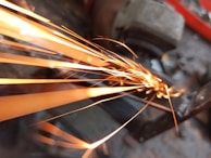 Bright, yellow-orange sparks fly from a piece of metal as it is being cut or grinded in a close-up view. The background shows a blurred setting with industrial tools and equipment.
