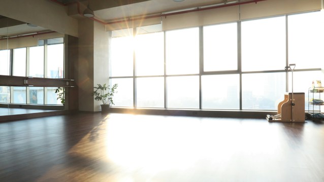 A spacious and sunlit room with a polished wooden floor, large windows allowing abundant natural light to stream in. A wall on the left has mirrors, and there's a potted plant near the mirror. On the right, near the windows, there is exercise equipment and a cart with various items.