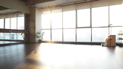 A spacious and sunlit room with a polished wooden floor, large windows allowing abundant natural light to stream in. A wall on the left has mirrors, and there's a potted plant near the mirror. On the right, near the windows, there is exercise equipment and a cart with various items.