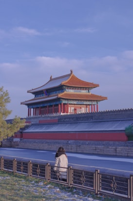 A traditional Chinese building with intricate architecture and vibrant colors stands prominently in the background. The roof is adorned with golden tiles, and red columns support the structure, reflecting classical Chinese design. In the foreground, a person in a white coat is walking along a pathway bordered by a decorative railing, with green grass and some fallen leaves visible.