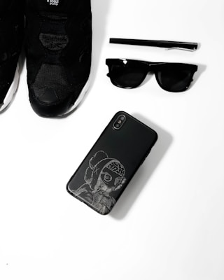 A collection of black fashion accessories including a pair of black sneakers, a black pen, black sunglasses, and a black smartphone with an artistic design on its case, arranged neatly on a white surface.