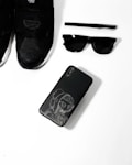 A collection of black fashion accessories including a pair of black sneakers, a black pen, black sunglasses, and a black smartphone with an artistic design on its case, arranged neatly on a white surface.
