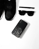A collection of black fashion accessories including a pair of black sneakers, a black pen, black sunglasses, and a black smartphone with an artistic design on its case, arranged neatly on a white surface.