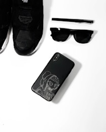 A collection of black fashion accessories including a pair of black sneakers, a black pen, black sunglasses, and a black smartphone with an artistic design on its case, arranged neatly on a white surface.