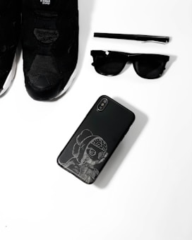 A collection of black fashion accessories including a pair of black sneakers, a black pen, black sunglasses, and a black smartphone with an artistic design on its case, arranged neatly on a white surface.