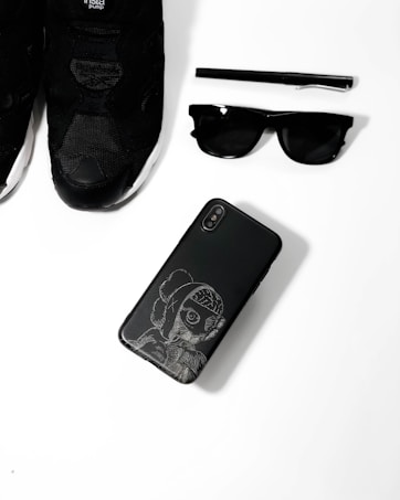 A collection of black fashion accessories including a pair of black sneakers, a black pen, black sunglasses, and a black smartphone with an artistic design on its case, arranged neatly on a white surface.