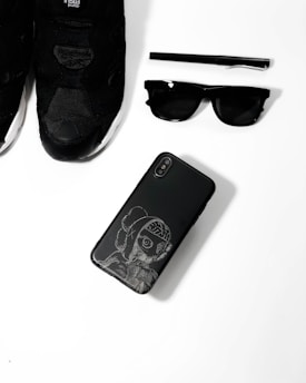 A collection of black fashion accessories including a pair of black sneakers, a black pen, black sunglasses, and a black smartphone with an artistic design on its case, arranged neatly on a white surface.