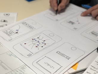 Sketches of mobile app designs are drawn on paper, showing multiple smartphone screen layouts with buttons and icons. A person is writing or drawing on one of the sketches using a pen. Additional notes and design elements are visible on the surrounding papers.