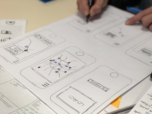 Sketches of mobile app designs are drawn on paper, showing multiple smartphone screen layouts with buttons and icons. A person is writing or drawing on one of the sketches using a pen. Additional notes and design elements are visible on the surrounding papers.
