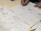 Sketches of mobile app designs are drawn on paper, showing multiple smartphone screen layouts with buttons and icons. A person is writing or drawing on one of the sketches using a pen. Additional notes and design elements are visible on the surrounding papers.