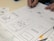 Sketches of mobile app designs are drawn on paper, showing multiple smartphone screen layouts with buttons and icons. A person is writing or drawing on one of the sketches using a pen. Additional notes and design elements are visible on the surrounding papers.