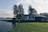 A serene landscape featuring a small house with a dark exterior, surrounded by lush green grass, next to a calm body of water. Tall trees stand near the house, and the sky is partly cloudy, providing a peaceful and tranquil setting.