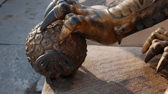A detailed bronze sculpture depicting a large, ornate claw grasping a intricately designed ball with a textured pattern. The craftsmanship highlights various geometric shapes and fine details, emphasizing a sense of power and artistry.