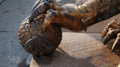 A detailed bronze sculpture depicting a large, ornate claw grasping a intricately designed ball with a textured pattern. The craftsmanship highlights various geometric shapes and fine details, emphasizing a sense of power and artistry.
