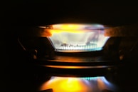 A close-up view of a gas stove burner with a bright blue flame, accented by hints of orange and yellow. The metal components of the burner are visible around the flame, and the surrounding area is dimly lit.