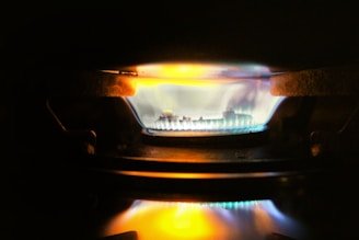 A close-up view of a gas stove burner with a bright blue flame, accented by hints of orange and yellow. The metal components of the burner are visible around the flame, and the surrounding area is dimly lit.