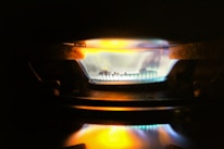 A close-up view of a gas stove burner with a bright blue flame, accented by hints of orange and yellow. The metal components of the burner are visible around the flame, and the surrounding area is dimly lit.