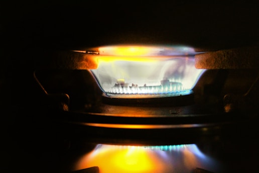 A close-up view of a gas stove burner with a bright blue flame, accented by hints of orange and yellow. The metal components of the burner are visible around the flame, and the surrounding area is dimly lit.